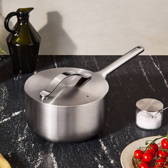 A Caraway Stainless Steel Sauce Pan, 3 qt, with 5-ply construction for even heating, rests on a dark marble countertop. Nearby are a green ceramic jug, a small salt container, and a white plate with cherry tomatoes, creating the warmth of an inviting kitchen.