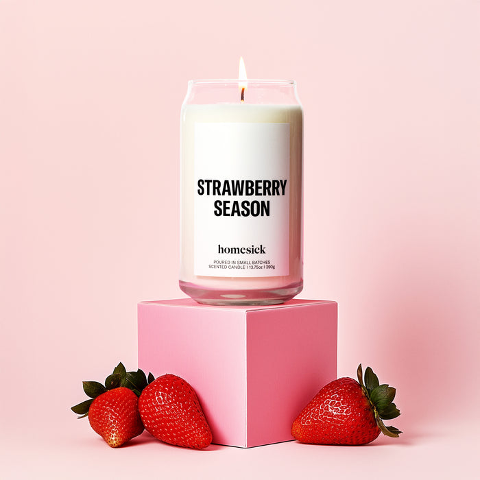 A Strawberry Season Candle by Homesick, made from cruelty-free soy wax, sits on a pink cube with three fresh strawberries in front, all set against a soft pink background, filling the space with natural scents.
