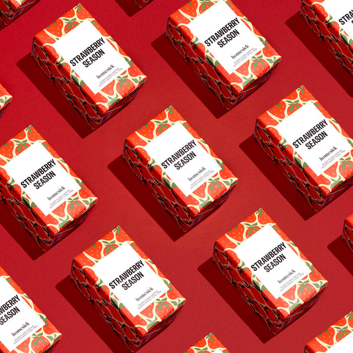 Homesicks Strawberry Season Candle boxes, featuring vibrant strawberry illustrations, are diagonally arranged on a red background, creating an appealing repetitive design inspired by the natural scents of soy wax candles.