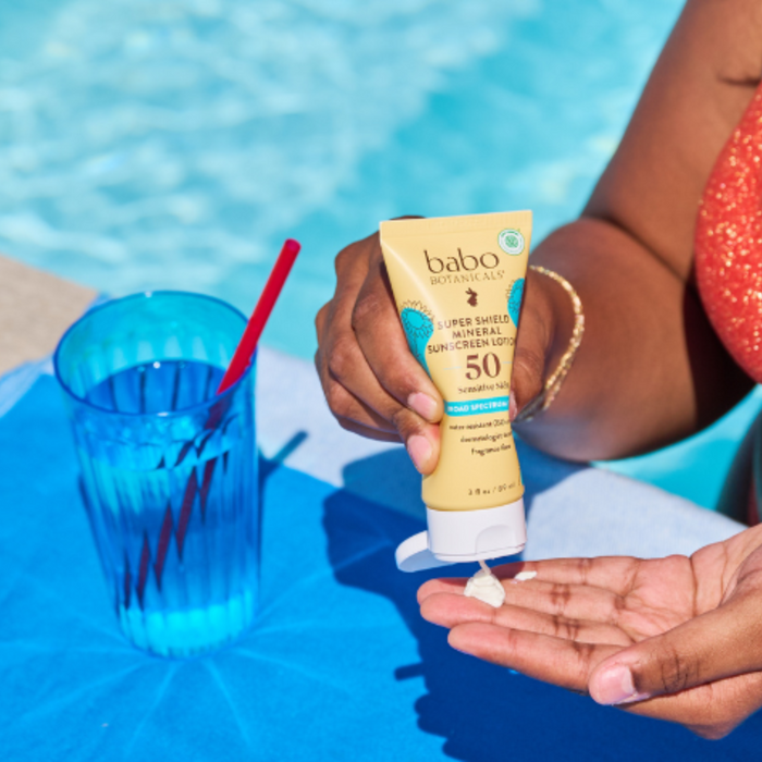 A person squeezes Babo Botanicals Super Shield Sunscreen Lotion SPF50 onto their hand beside a blue cup with a red straw by the pool, offering perfect protection for sensitive skin.