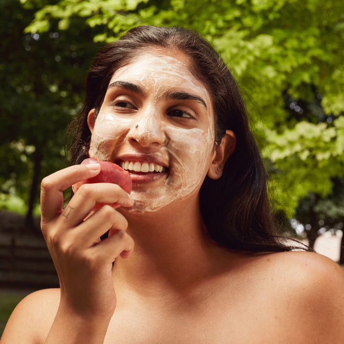 A woman with long dark hair and a face mask smiles, holding a peach outdoors—her radiant skin might be from the Three Ships Superfruit Lactic + Multifruit 8% AHA Exfoliating Mask, packed with fruit-based AHAs for glowing results.