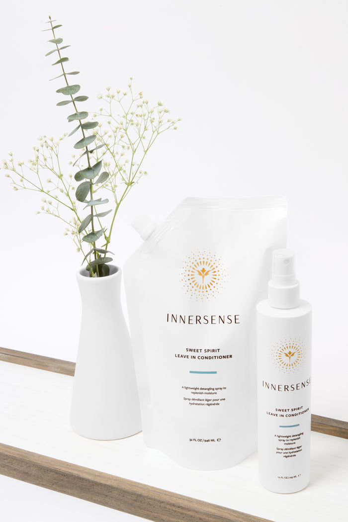 A white vase with green foliage and babys breath sits beside Innersense Organic Beauty Sweet Spirit Leave In Conditioner in a white refill pouch and spray bottle, displayed on a white surface with a wooden edge.
