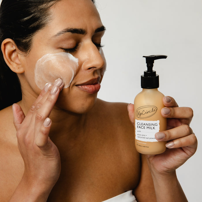 Smiling, a person applies The Discovery Set to their cheek, enjoying the transformative effects of a starter skincare routine. They hold an UpCircle product against a plain white background, highlighting the upcycled beauty brand.
