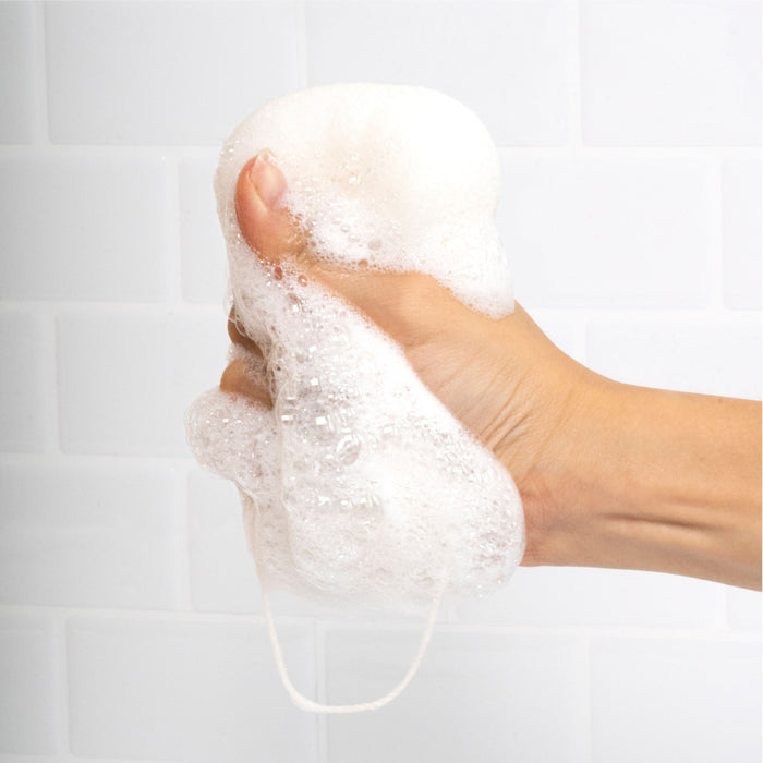 A hand tightly squeezes The Konjac Body Sponge by Everist, made from natural konjac plant fibre, creating rich foam and bubbles against a white tiled background.