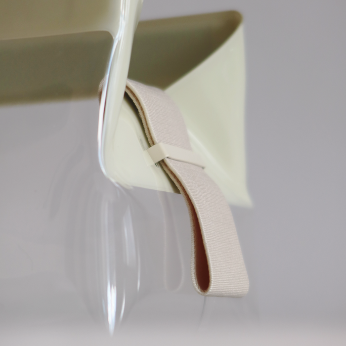 A close-up of The Parcel by Cadence, a TSA-approved travel bag, shows its clear curved surface with a beige elastic band looped through a small plastic clip, set against a light gray background.