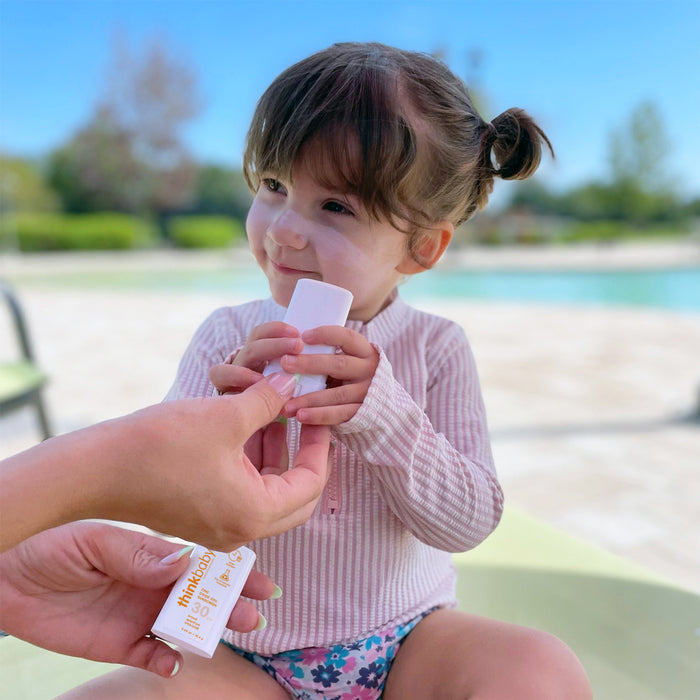 A young child in a striped shirt applies thinksun thinkbaby SPF 30 Face & Body Sunscreen Stick to her face by a pool on a sunny day, as an adult helps hold the sunscreen container.