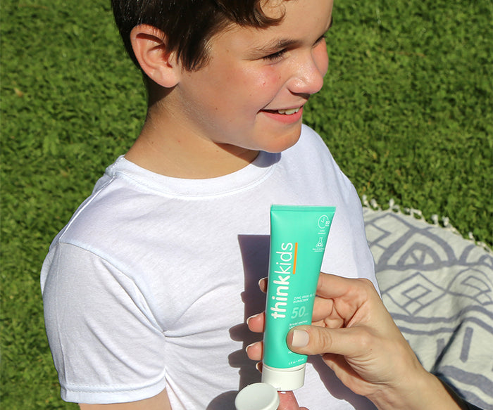 A child smiles outdoors while someone applies thinksun thinkkids SPF 50 Sunscreen, a mineral-based zinc formula, to their arm. Grass and a patterned blanket can be seen in the background.