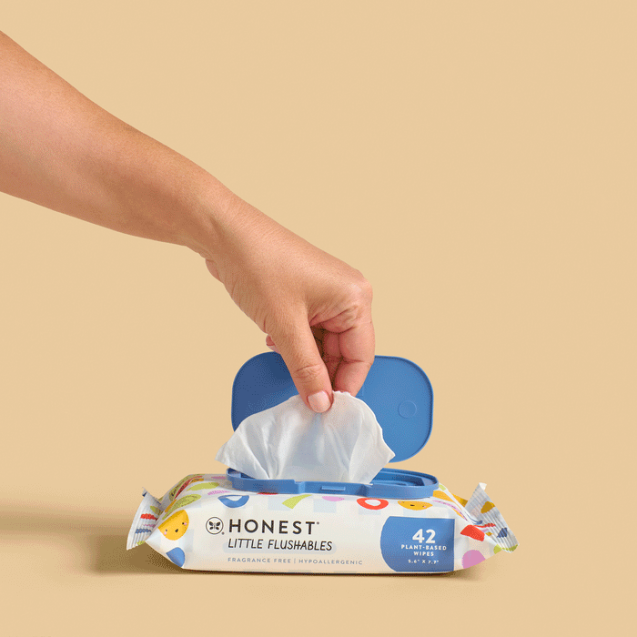 A hand pulls a wipe from a package of Toddler Flushable Wipes Designed for Potty Training by The Honest Company. The packaging features playful designs and emphasizes its fragrance-free, hypoallergenic qualities, with an open blue lid on top.