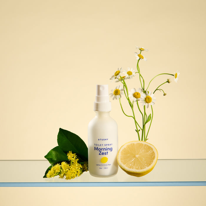 A white TUSHY Toilet Spray bottle sits on a glass shelf with yellow flowers, green leaves, a half lemon, and daisies—perfect for eliminating odors and brightening any space.