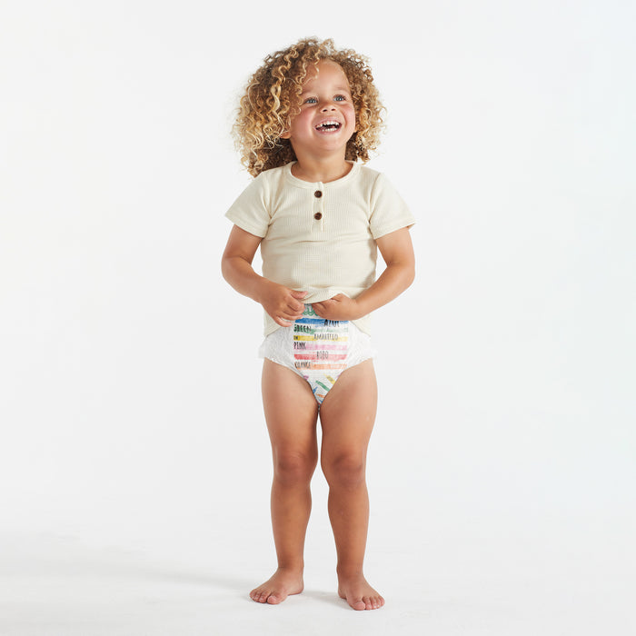 A young child with curly hair is smiling broadly in a beige short-sleeved shirt and The Honest Companys colorful Training Pants - 2T/3T, against a plain white background.