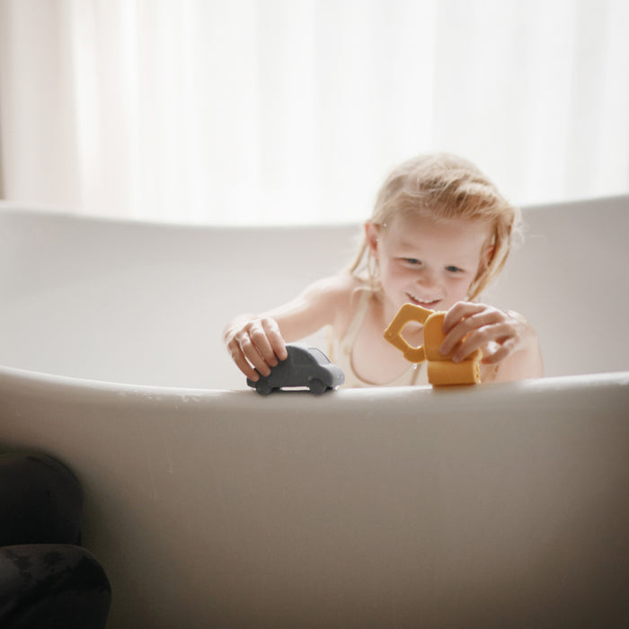 A young child with blonde hair plays with the mushie Vehicles Mold-Free Bath Toy Set – Soft Silicone while sitting in a white bathtub, as soft natural light filters in from a window behind.