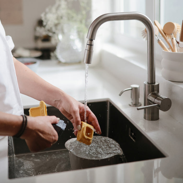 A person washes a metal pot while utensils and mushies Vehicles Mold-Free Bath Toy Set – Soft Silicone, made of food grade silicone for safe, easy cleaning, sit on the counter near the sink.