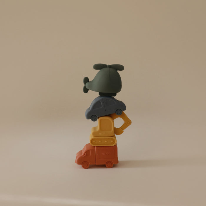 The mushie Vehicles Mold-Free Bath Toy Set features a stack of minimalist, colorful vehicles—a green helicopter, blue car, yellow bulldozer, and orange bus—made from soft food-grade silicone and set against a plain beige background.