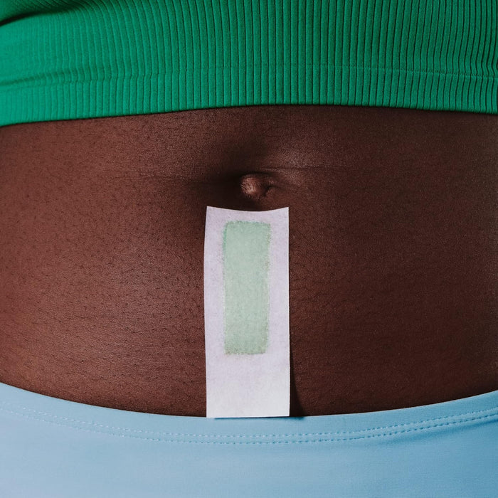 A person with a medium skin tone in a green top and light blue pants uses the billie Wax Kit, applying a rectangular strip to their lower stomach below the belly button. After hair removal, they use soothing serum wipes to calm their skin.