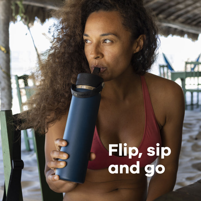 A person with long curly hair in a red bikini top sips from a Hydro Flask Wide Mouth Bottle with Flex Straw Cap under thatched roof. Sand and wooden furniture are visible in the background. Text reads, Flip, sip and go.