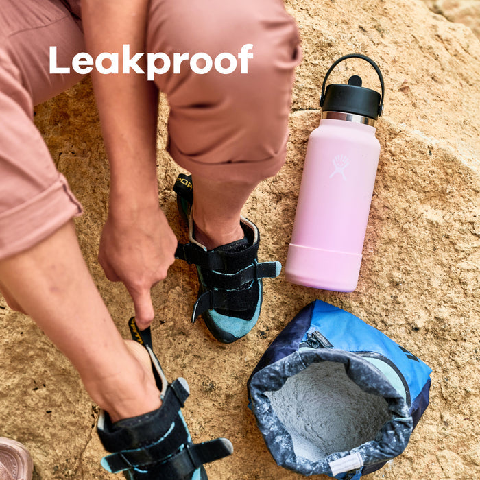 A person in pink pants slips on a black climbing shoe against a sandy backdrop. Nearby, a pink Hydro Flask Wide Mouth Bottle with Flex Straw Cap sits next to a blue chalk bag. The word Leakproof is prominently displayed in the top left corner.