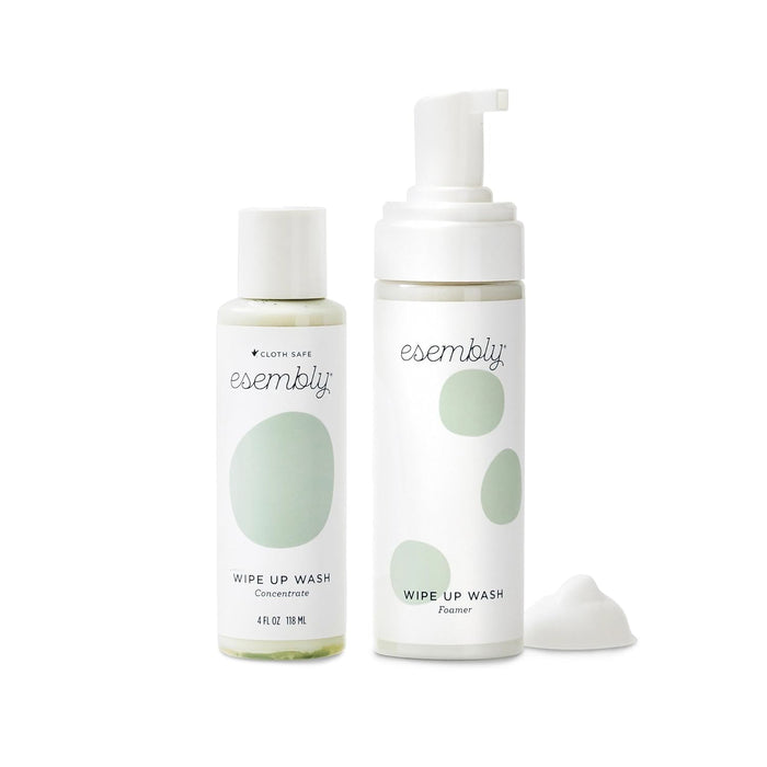 The Wipe-Up Wash Kit by Esembly Baby features two bottles: a 4 oz concentrate with a flip cap and an organic foaming wipes cleanser in a pump bottle. Both have minimalist designs with green abstract shapes, and foam is displayed in front of the pump bottle.