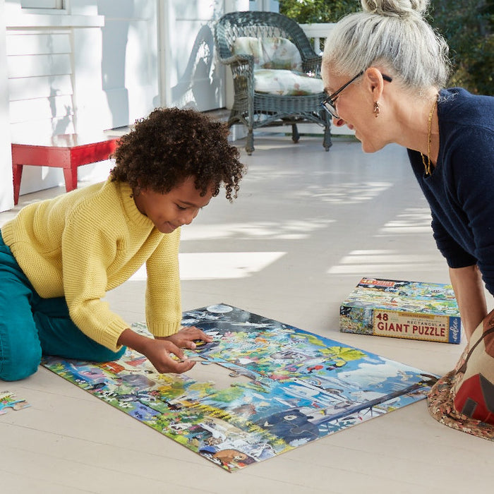 A child and an older woman smile together on a porch as they assemble the colorful eeBoo Piece & Love Within the Biomes 48 Piece Giant Puzzle. The box and loose pieces are nearby, lit by sunlight streaming onto the wooden floor.