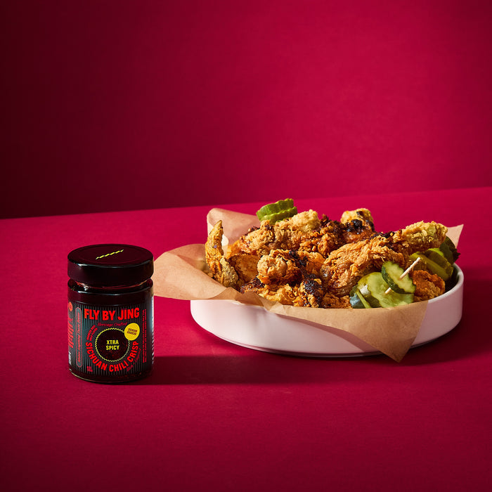A plate of fried chicken with pickles sits on parchment paper beside a jar of Fly By Jing Xtra Spicy Sichuan Chili Crisp, made with extra-hot Chinese chilis, set against a solid red background.